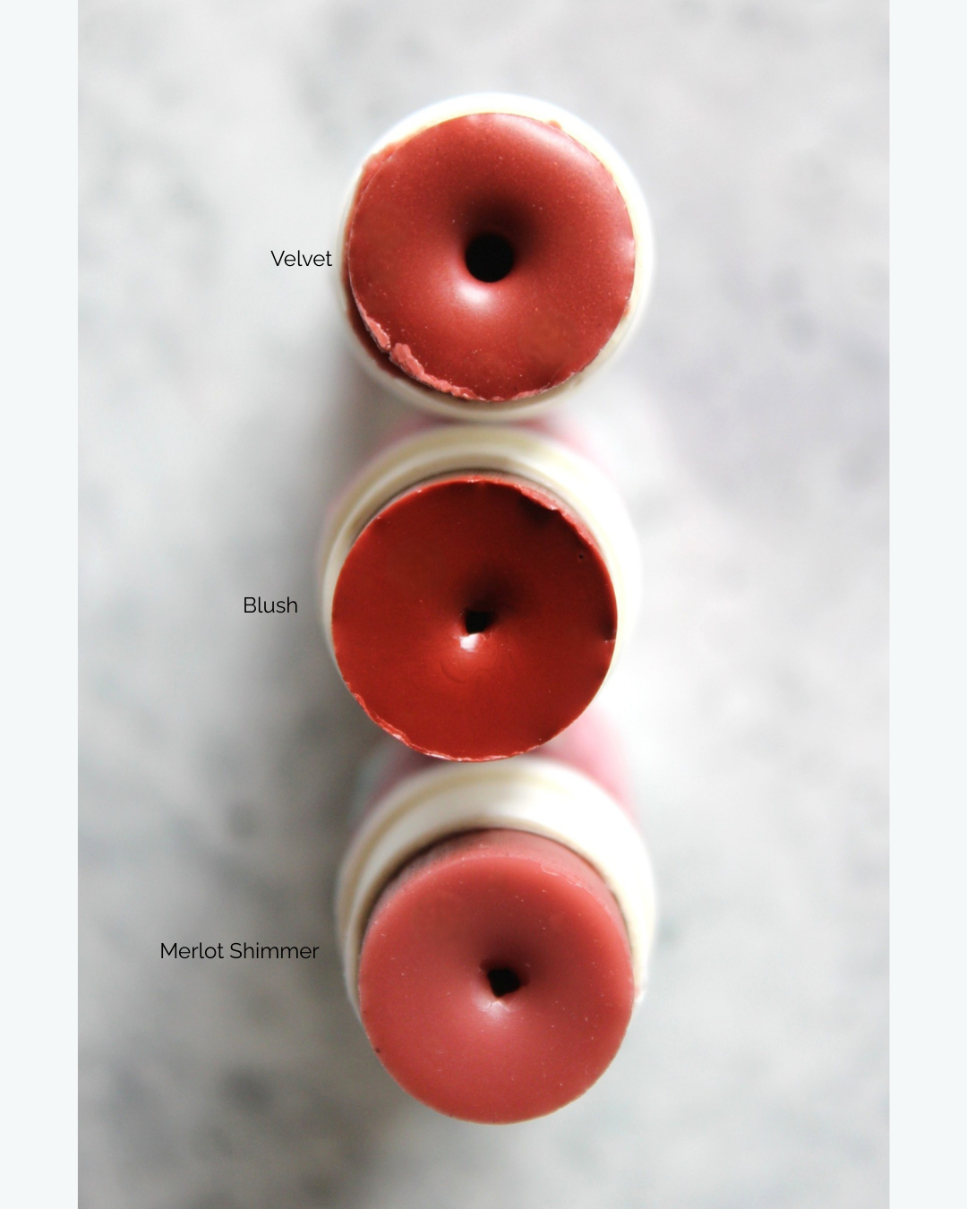 Three red lip products labeled 'Velvet', 'Blush', and 'Merlot Shimmer' on a light gray background.