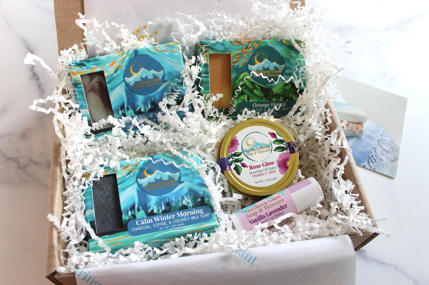 Gift box with bath and body products including soaps and skincare on a marble surface.