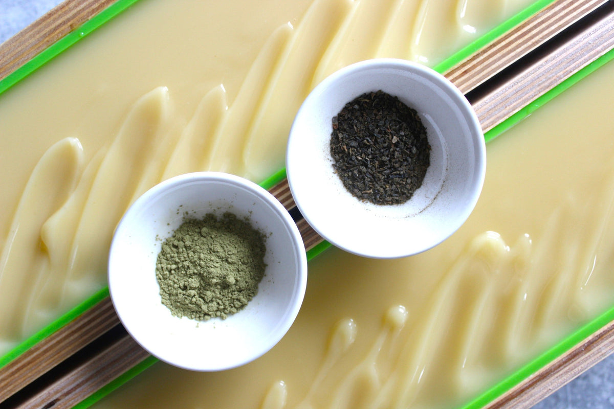 Two small white bowls, one with matcha powder, the other with Alaska sea kelp, sitting on two wooden soap molds containing eucalyptus and Alaska sea kelp soap.