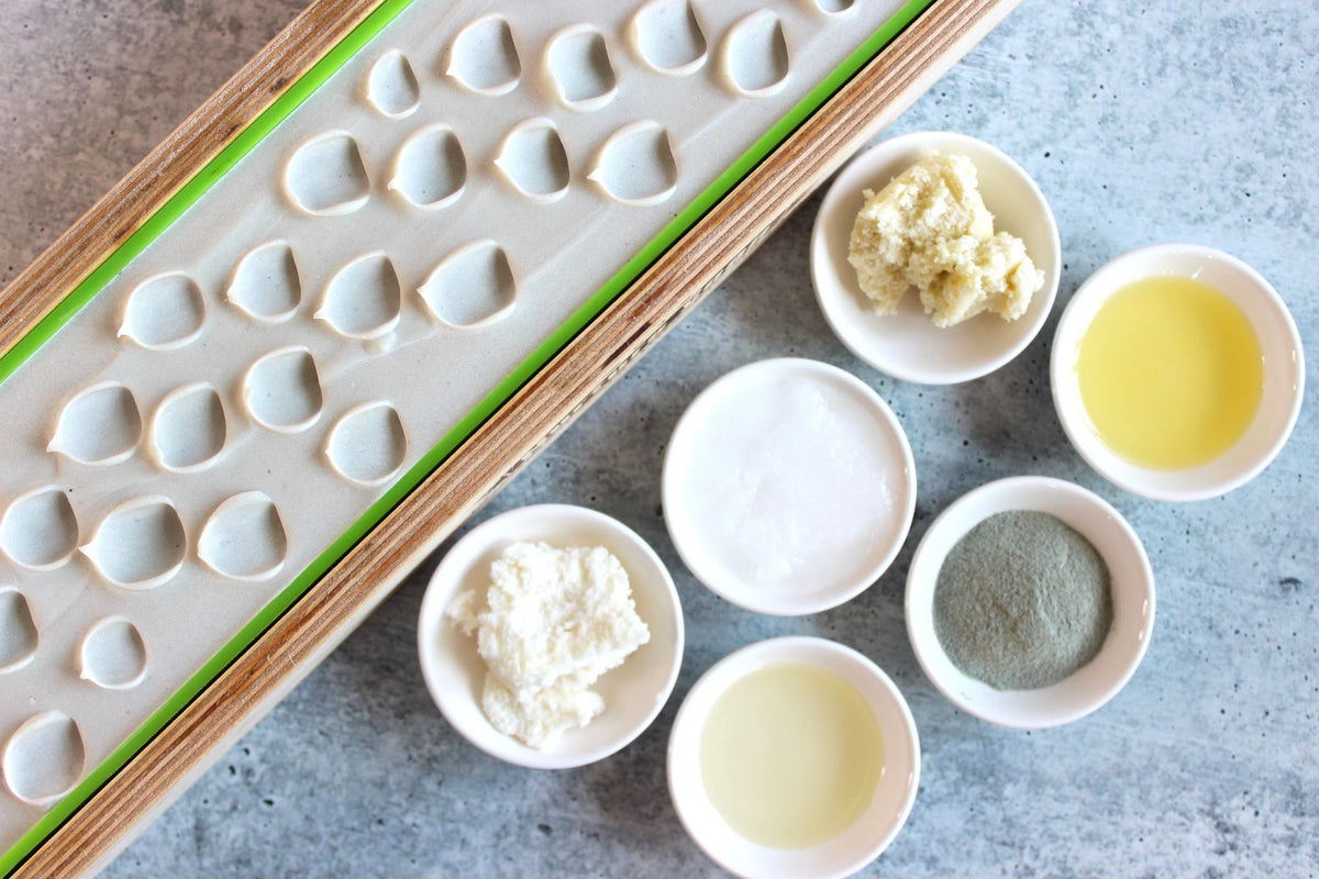 An overhead flat-lay of a handmade soap-making ingredients, and a loaf of soap in a wooden mold. Several small white bowls containing ingredients like salt, kokum butter and clay on a gray stone surface.