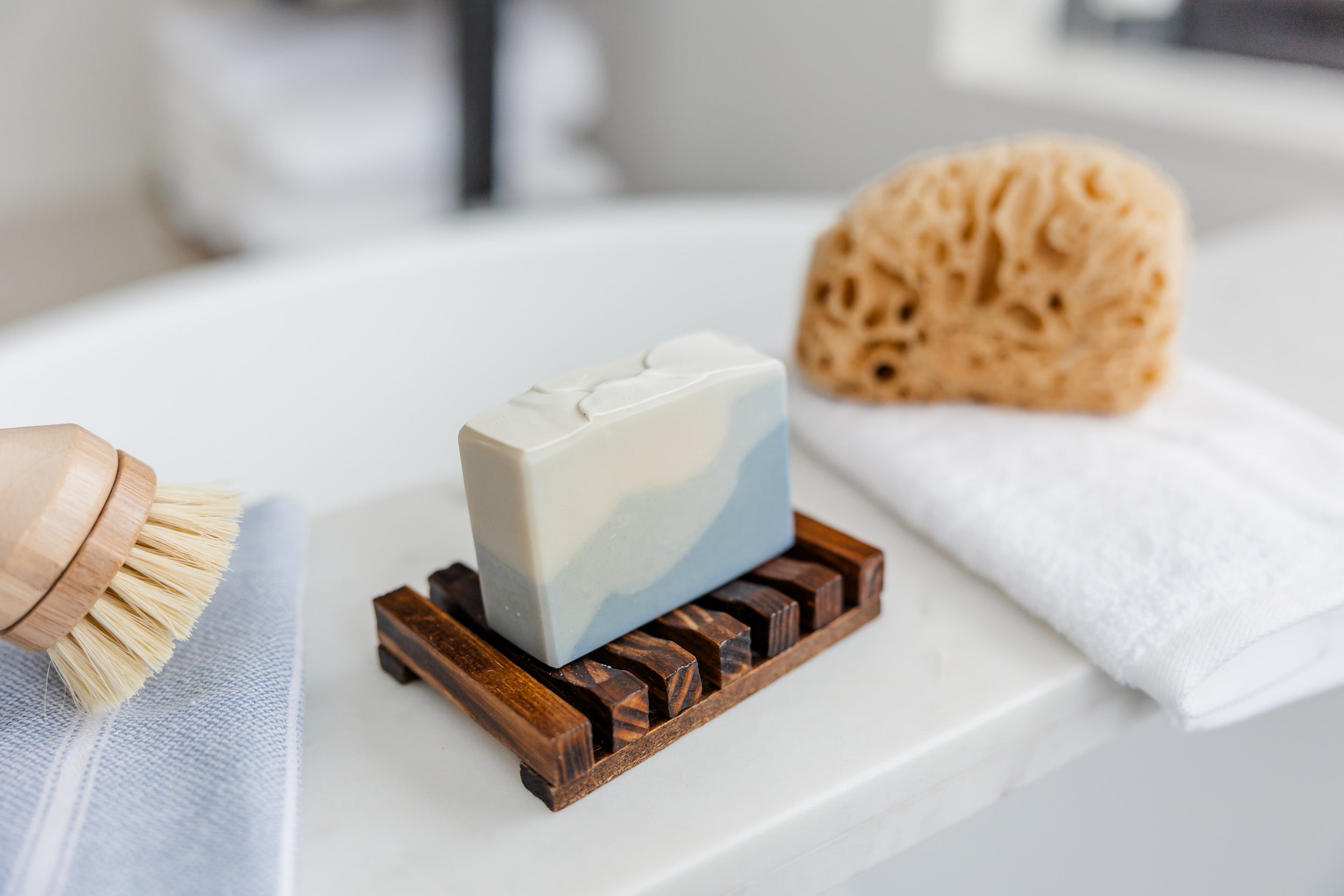 Bar of soap on a wooden soap dish with a sponge and brush on a white surface.