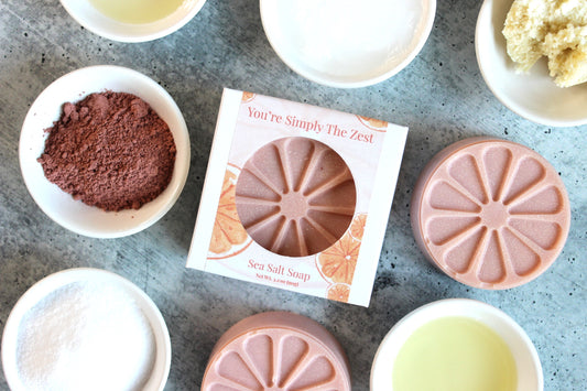 Pink Grapefruit Soap called You're Simply the Best is shaped like a slice of citrus.  Pictured in pink with ingredients in white bowls all around the soap.
