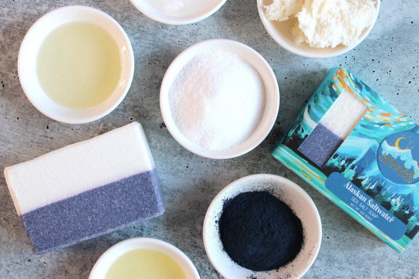 Alaskan Saltwater soap featured in a blue box with mountains as well as ingredients used in the soap in little white bowls.