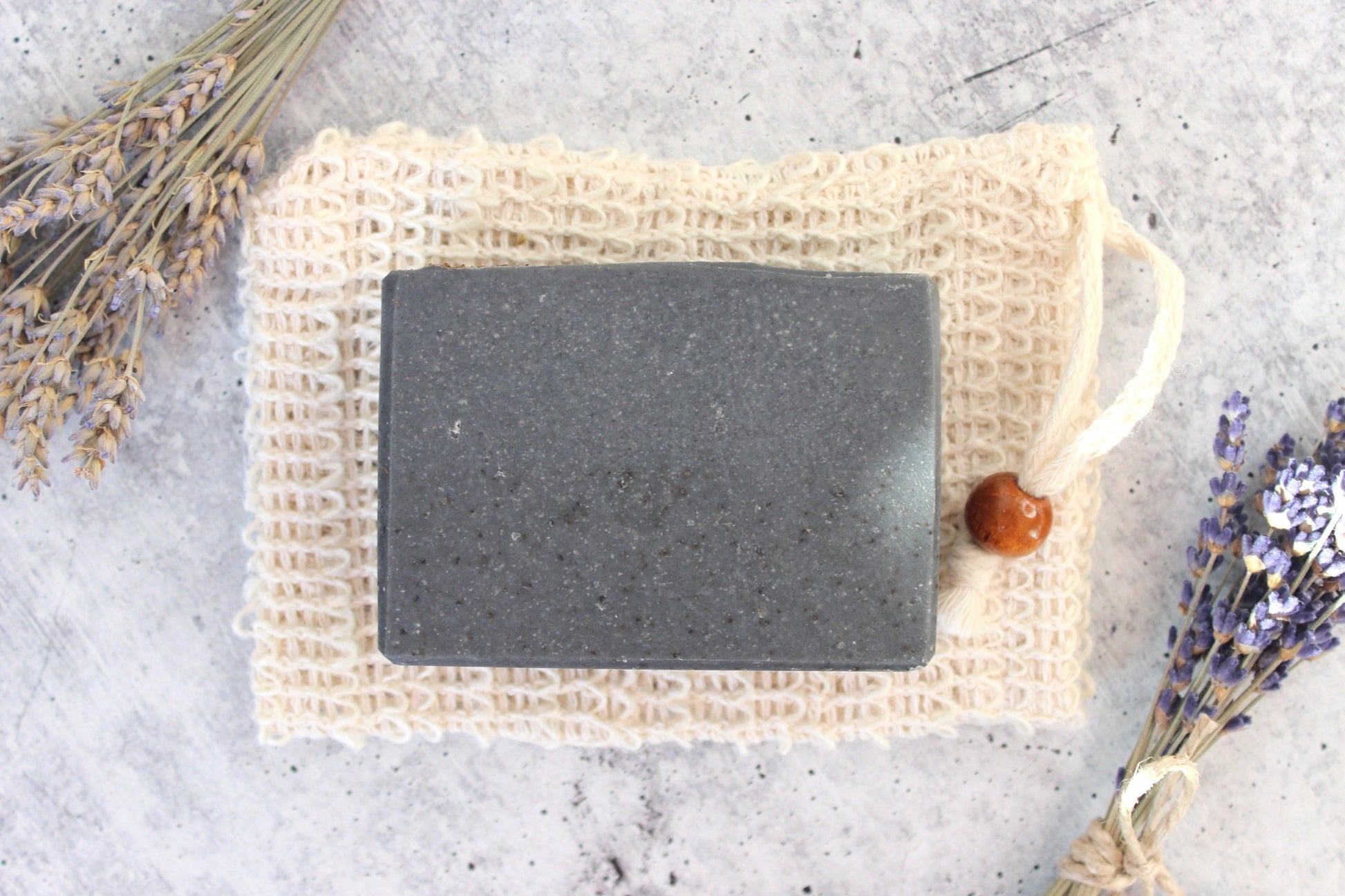 Black soap bar on a natural fiber soap bag with lavender on a gray background