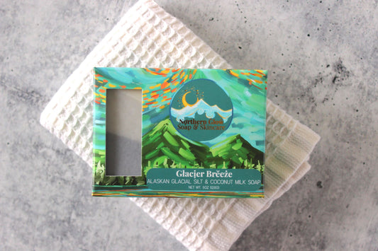 Charcoal soap with glacier silt. Pictured in a green box with mountains. Sitting on a white wash cloth.