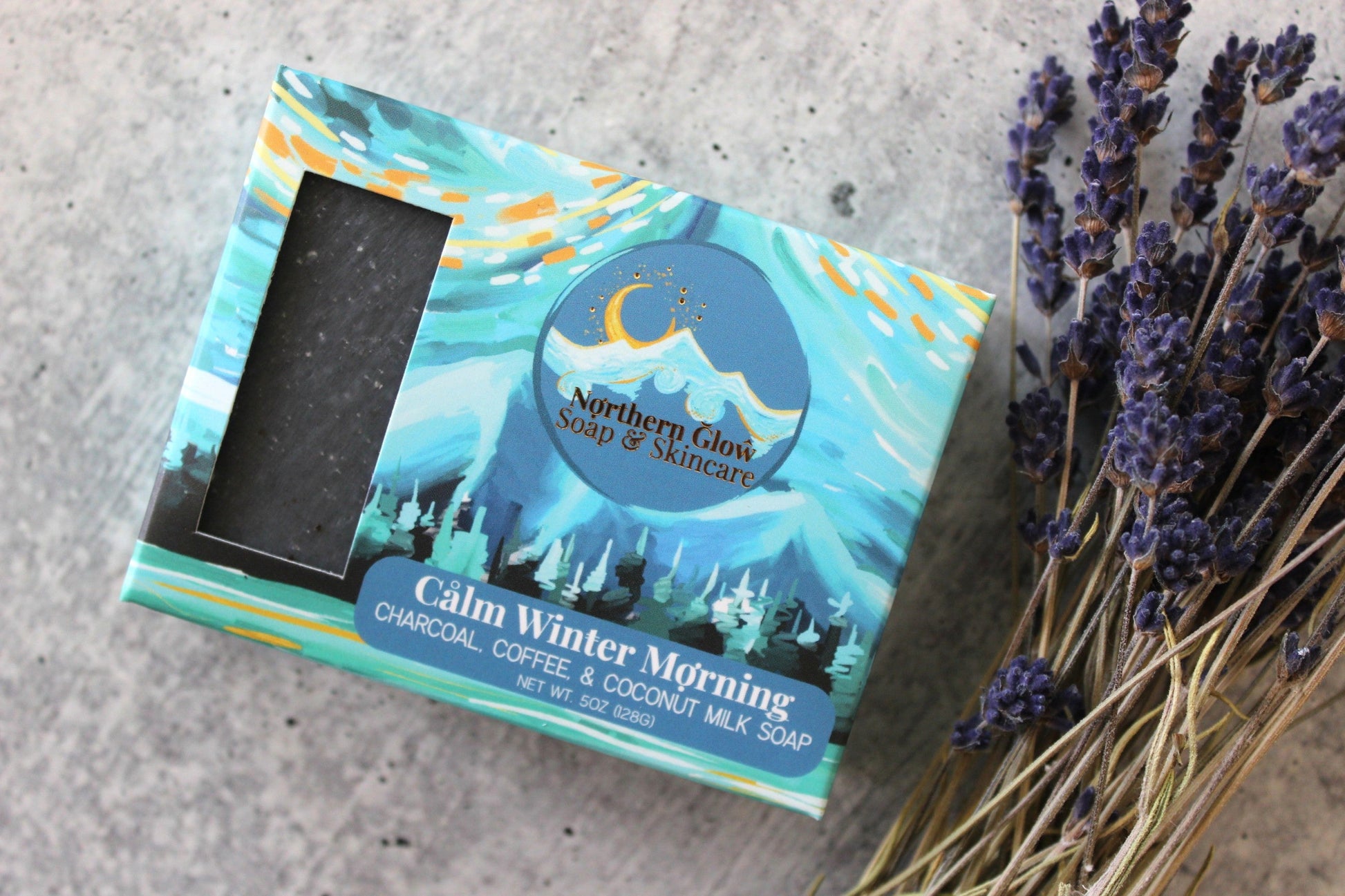 Dark gray soap in a blue box, next to dried lavender, on a concrete background.