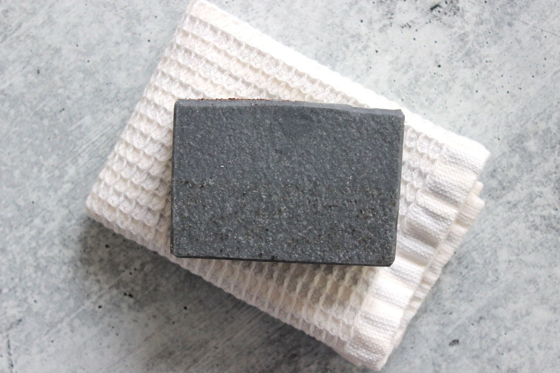 Bar of charcoal soap on a textured white towel against a gray concrete background