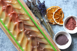 Lavender goat milk soap in a wooden mold.  Dried lavender, dried orange slices next to purple and pink clay.