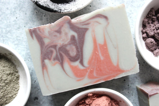 Gray soap with swirls of purple and pink next to white bowls with clay and charcoal.
