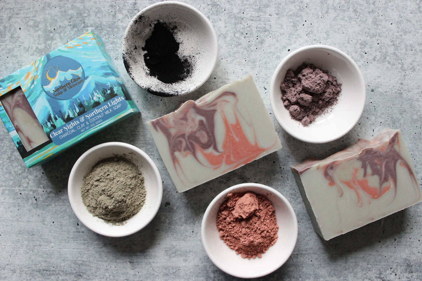 Gray soap with swirls of pinks and purples, next to a blue soap box.  Bowls of green, pink, and purple clay.