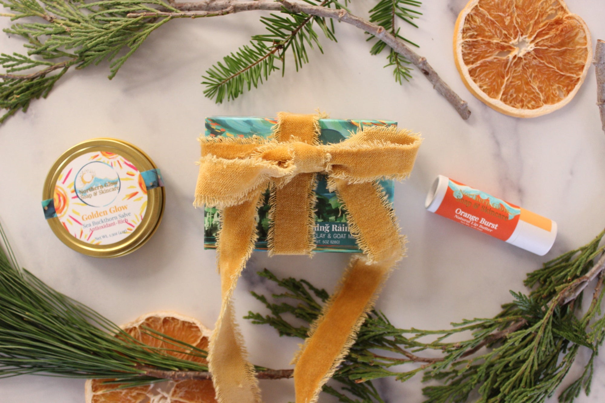 Natural skincare products (a salve, soap, and lip balm) surrounded by sprigs of pine, cedarwood, and dried orange slices on a white marble surface.