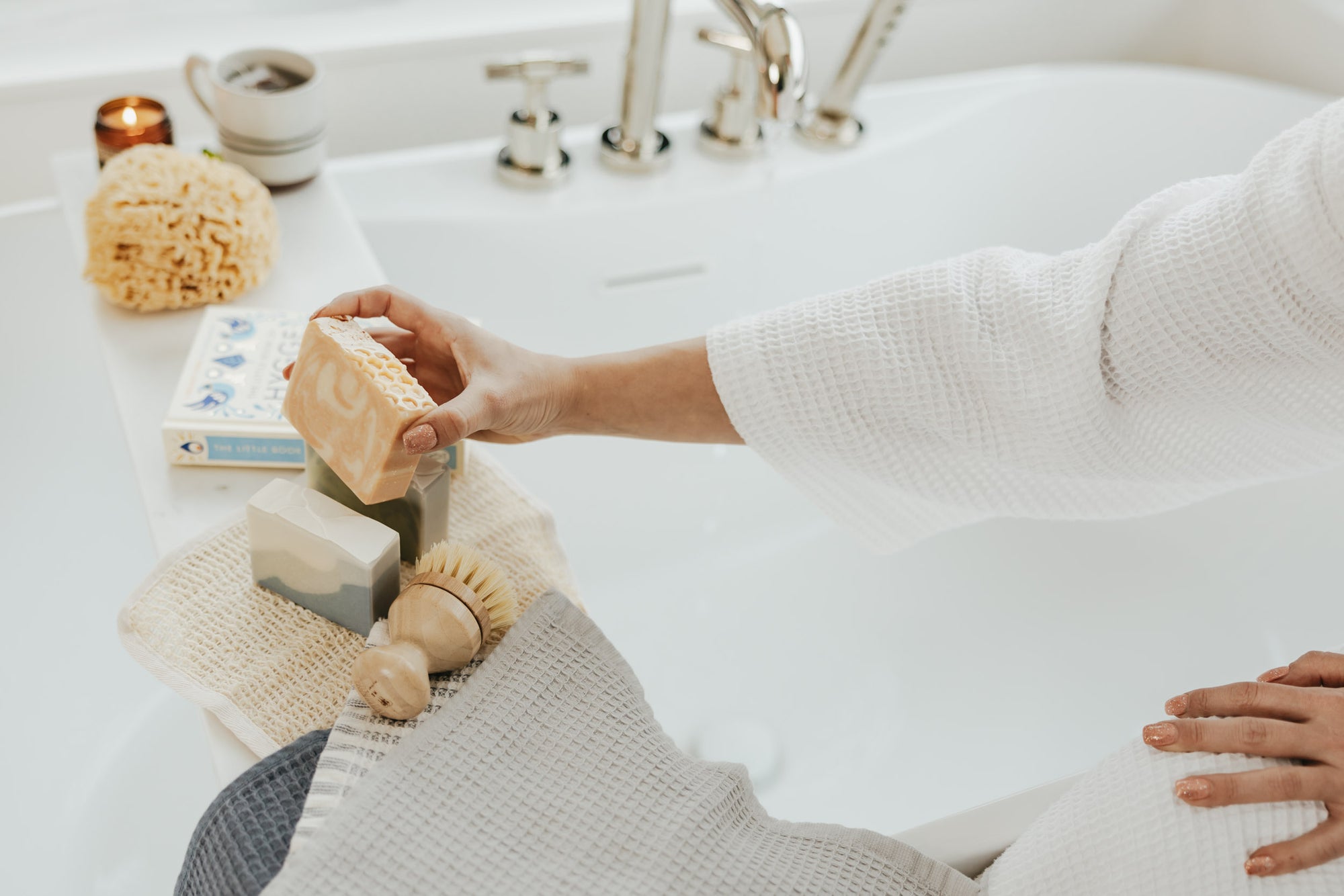 A woman grabbing a bar of soap with a bathtub and candle glowing in the background.