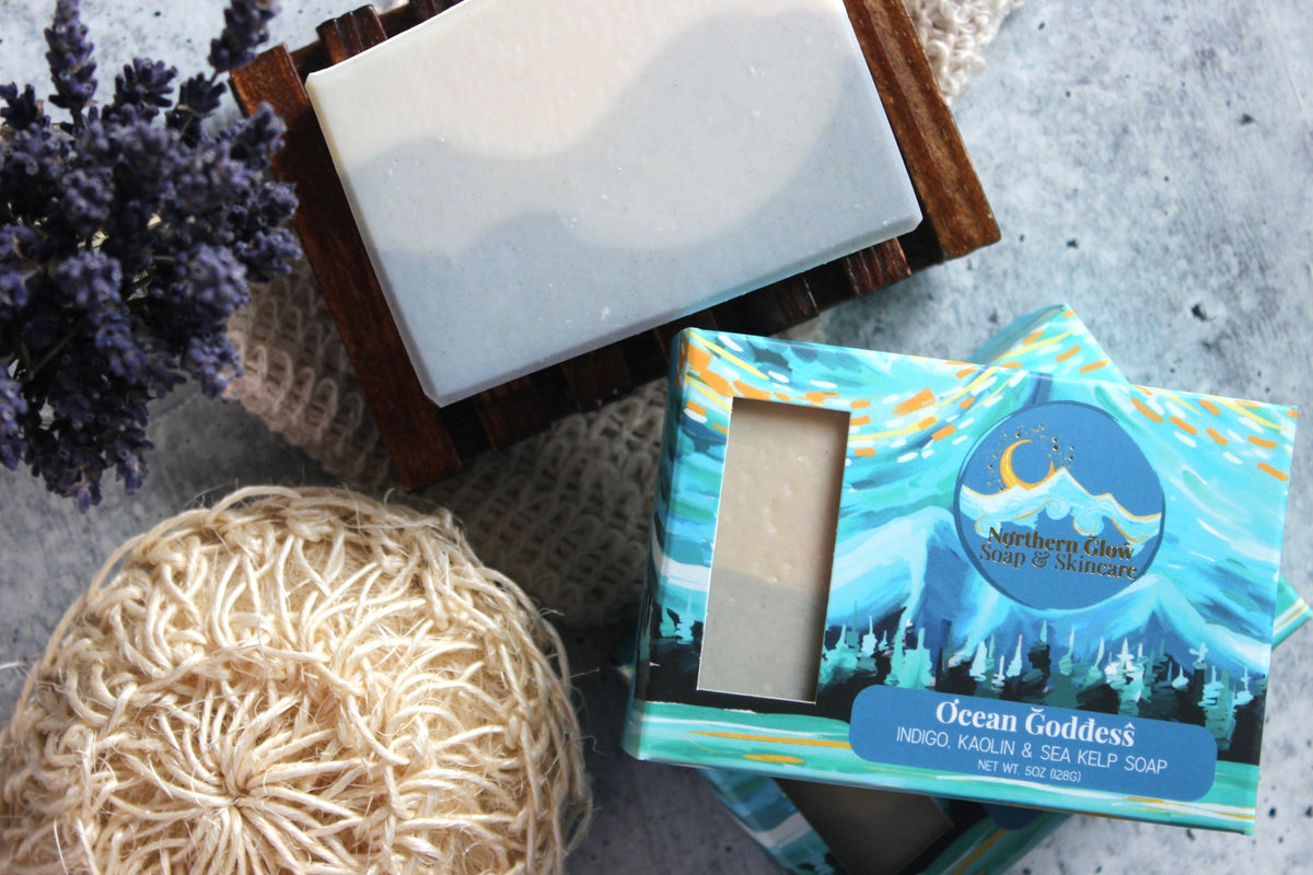 A boxed bar of Ocean Goddess soap and the soap itself displayed on a bamboo soap dish, next to a natural loofah.  The box reads 'Northern Glow Soap & Skincare Ocean Goddess' and shows a blue illustrated mountain and river design.