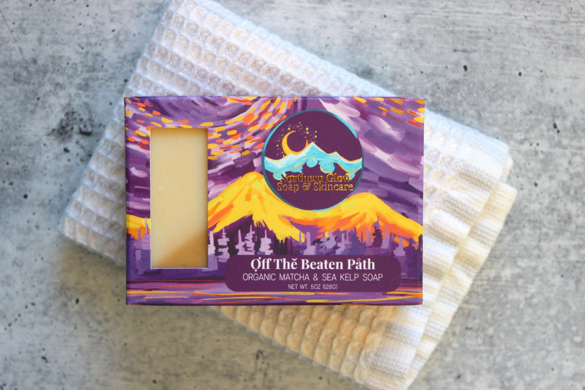 Eucalyptus and Alaska sea kelp bar soap in a purple box with golden colored mountains, resting on a white washcloth and concrete counter.