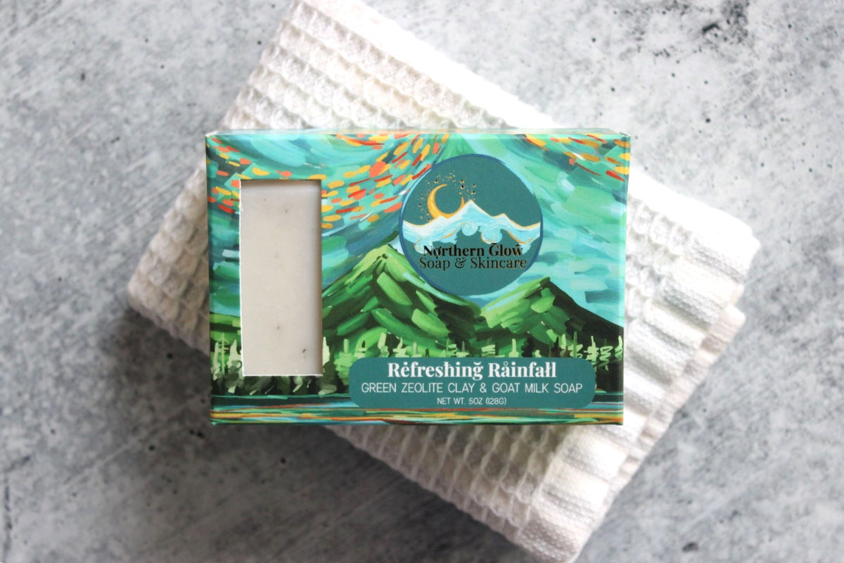 A packaged bar of soap from Northern Glow Soap & Skincare, labeled 'Refreshing Rainfall,' placed on top of a folded white textured towel against a gray stone-like surface.