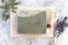Load image into Gallery viewer, Alpine Rainstorm Soap, made with bergamot, cedarwood and goat milk, resting on a cambric soap saver.  Dried peppermint and lavender flowers surround the soap.
